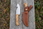 Thicket Warden satin D2 gut hook hunting knife with olive wood handle and brown leather sheath on weathered decking