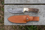 Royal Stag Damascus gut hook hunting knife with stag antler handle, brass bolster and tan leather sheath on a weathered board