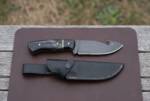 Blackthorn satin D2 gut hook hunting knife with black buffalo horn handle and black leather sheath on a leather mat