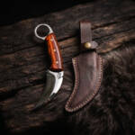 Copper Crescent mirror polished D2 karambit knife with rosewood handle and dark brown leather sheath on dark wood