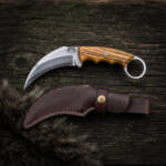 Smokeclaw hand forged high carbon karambit knife with olive wood handle and dark brown leather sheath on rustic wood