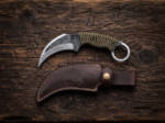 Bush Viper hand forged high carbon karambit knife with olive green paracord handle and dark brown leather sheath on rustic wood