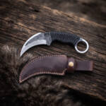 Black Talon hand forged high carbon karambit knife with black paracord handle and oxblood leather sheath on dark wood