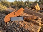 Mesa Drifter satin carbon steel cowboy bull cutter knife with walnut burl handle and tooled leather sheath on mesquite logs at sunset