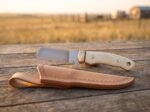 Pale Longhorn satin D2 cowboy bull cutter knife with camel bone handle, brass bolster and tooled leather sheath on a corral rail at sunset