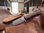 Latigo Ranger satin D2 cowboy bull cutter knife with walnut handle and leather sheath resting on a western saddle