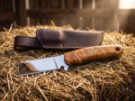 Chrome Wrangler mirror polished D2 cowboy bull cutter knife with olive burl handle and brown leather sheath on a hay bale