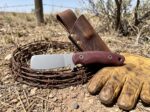 Oxblood Rider stonewashed carbon steel cowboy bull cutter knife with burgundy Micarta handle and leather sheath beside barbed wire on a ranch