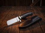 Night Herder mirror polished D2 cowboy bull cutter knife with ebony wood handle and black leather sheath on dark barn wood