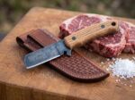 Bluestem Ridge acid etched spring steel cowboy bull cutter knife with oak handle and tooled leather sheath beside ribeye steaks on a butcher block