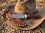 Lariat Rose satin D2 cowboy bull cutter knife with rosewood handle, brass bolster and leather sheath resting on a felt cowboy hat