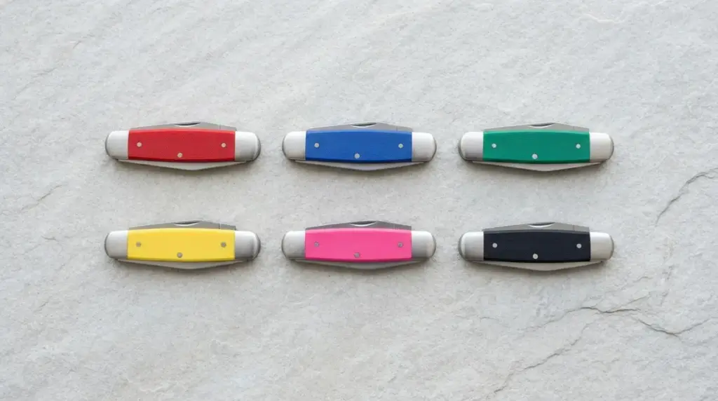 Six identical small knives with red, blue, green, yellow, pink and black handles on grey stone