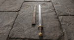 Sea Wolf D2 steel Viking sword with straight double edged fullered blade, brown leather wrapped grip, brass guard and brazil-nut pommel beside its leather wrapped wooden scabbard on stone