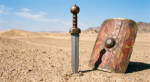 Bronze Legion D2 steel Roman gladius short sword with broad leaf shaped bead blasted blade, ribbed hardwood grip and spherical bronze pommel, standing in desert sand beside a scutum shield